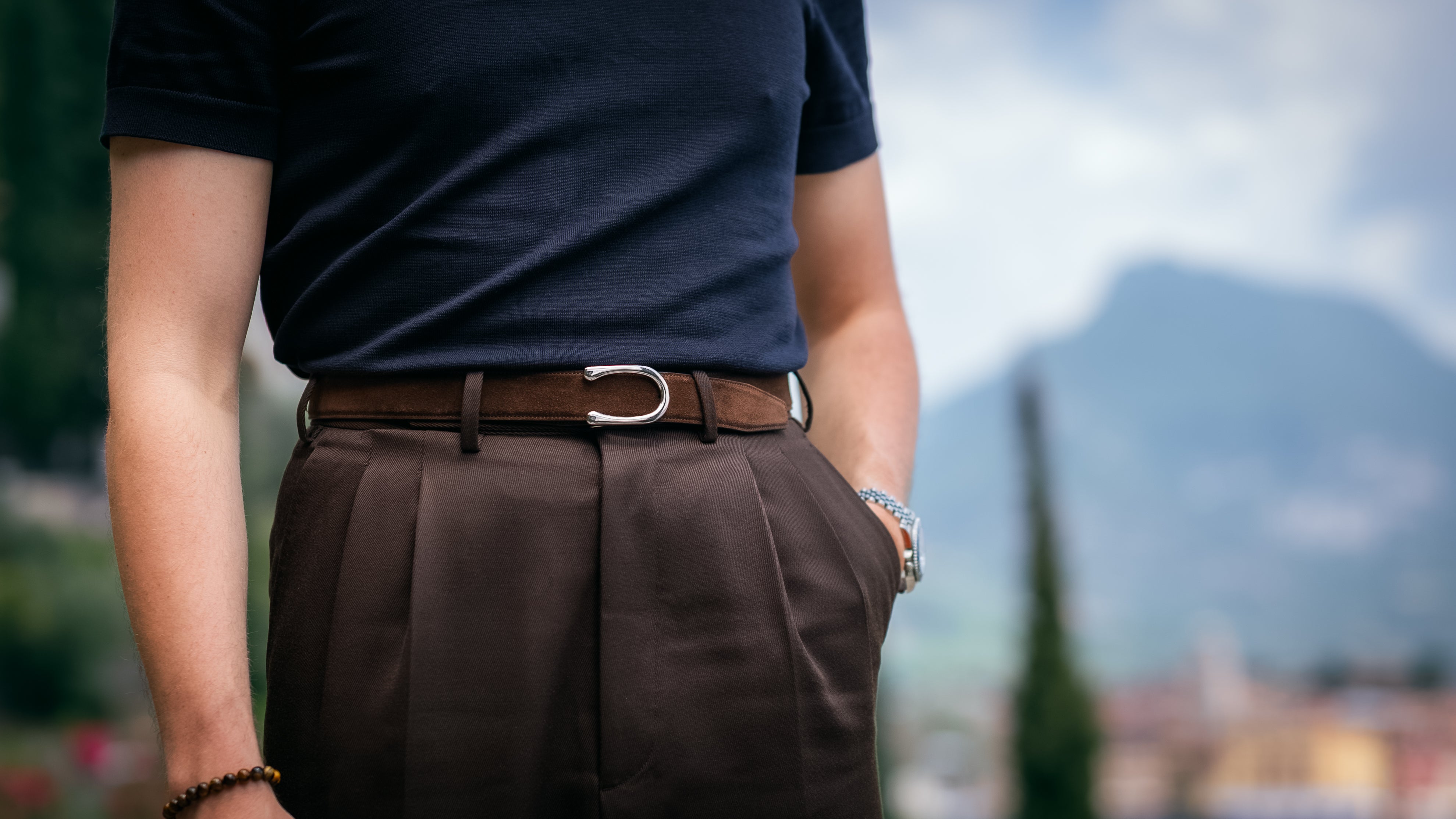 Person wearing a navy shirt and brown pants with a Goldfels Signature Belt in Havana Brown Suede and a Model 2 Palladium Buckle in front of a blurred Italian scenic background.