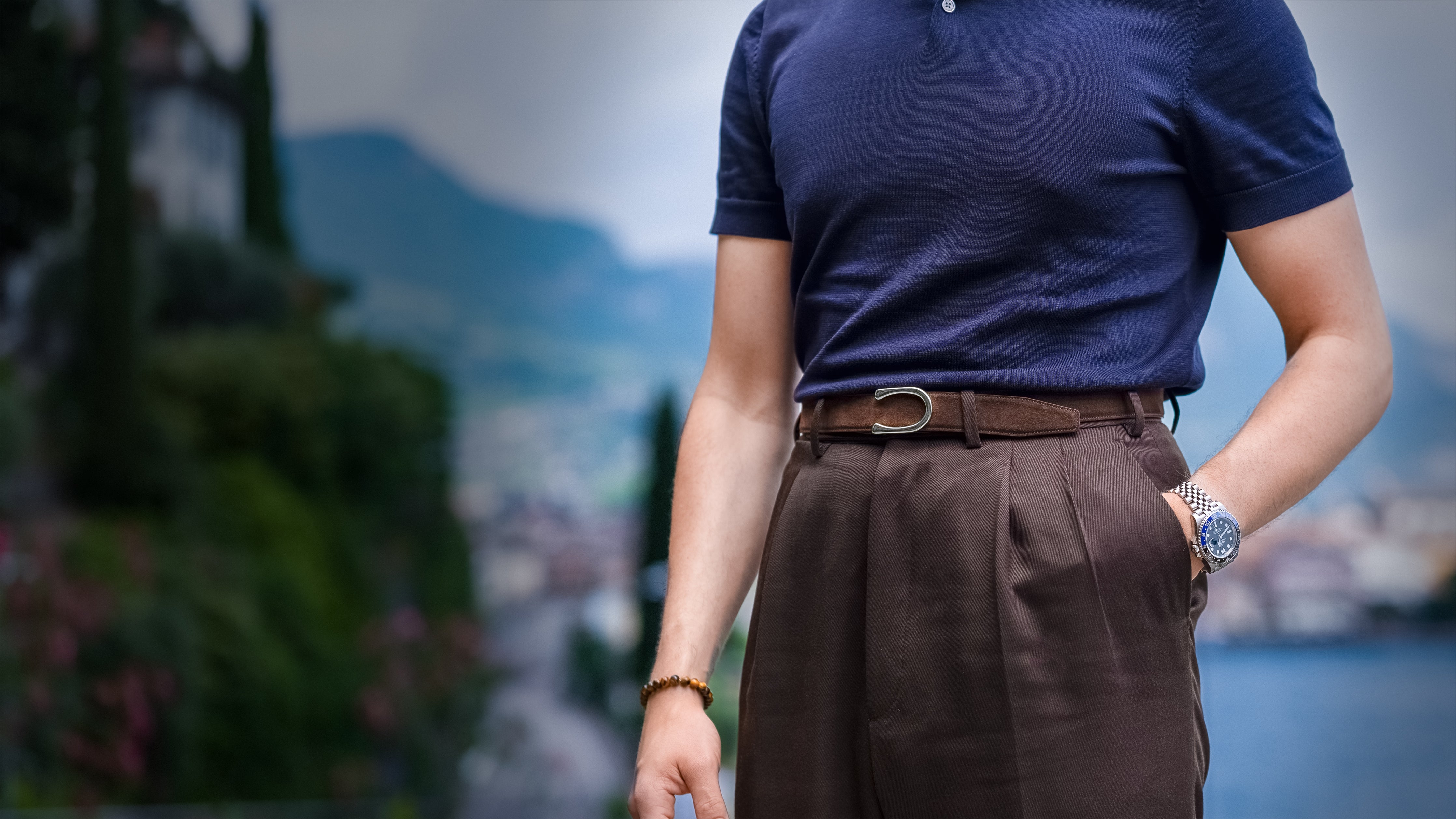 Gentleman wears a Havana Brown Goldfels Suede Belt with Palladium Buckle Model 2 paired with elegant brown trousers and a luxurious navy knitted polo shirt in front of a blurred Italian landscape.